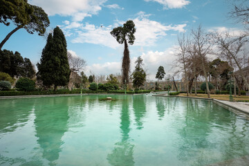 The serene pools of the Golestan Palace are mirrored by towering trees against a cloudy sky....