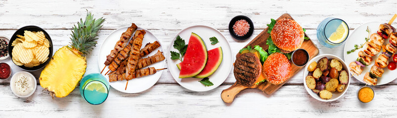 Summer BBQ food table scene. Hamburgers, meat skewers, potatoes, fruit and snacks. Top down view on a white wood banner background.
