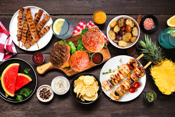 Fototapeta premium Summer BBQ food table scene. Hamburgers, meat skewers, potatoes, fruit and snacks. Top view on a dark wood background.