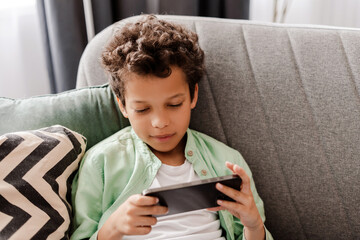 Excited African American boy lying holding mobile phone, playing games, social media