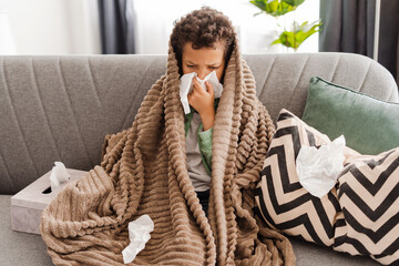 Little upset African American boy has a cold, holding napkin, covered with blanket