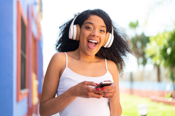 Young African American woman with headphones at outdoors surprised and sending a message