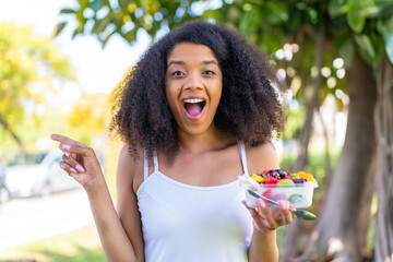 Young African American woman holding a bowl of fruit at outdoors surprised and pointing finger to...