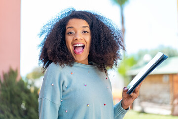 Young African American woman holding a tablet at outdoors with surprise and shocked facial...
