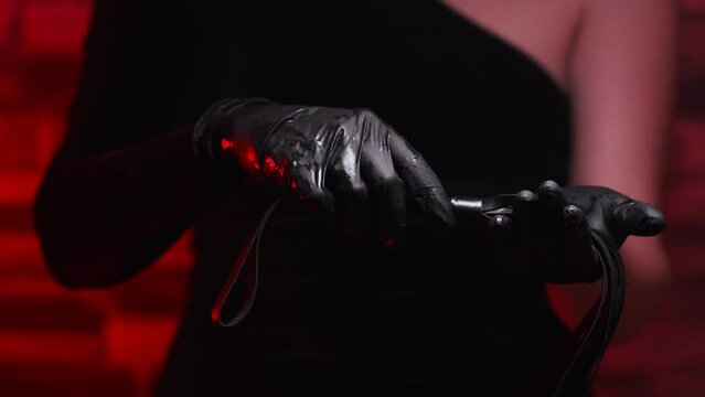 Close-up of female hands in black gloves holding a BDSM whip on a red background. BDSM is an outfit for adult sex games. Sex shop Adult store
