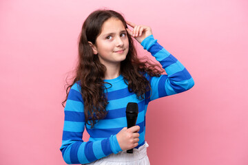 Little singer girl picking up a microphone isolated on pink background having doubts and with...