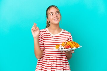 Little caucasian girl holding waffles isolated on blue background with fingers crossing and wishing...