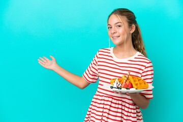 Little caucasian girl holding waffles isolated on blue background extending hands to the side for...