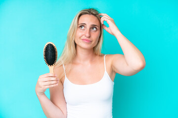 Young caucasian woman with hair comb isolated on blue background having doubts and with confuse...