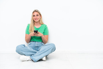 Fototapeta premium Young caucasian woman sitting on the floor isolated on white background sending a message with the mobile