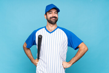 Young man playing baseball over isolated blue background posing with arms at hip and smiling