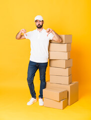 Full-length shot of delivery man among boxes over isolated yellow background showing thumb down