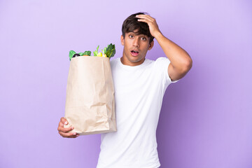 Young man holding a grocery shopping bag isolated on purple background doing surprise gesture while...