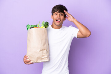 Young man holding a grocery shopping bag isolated on purple background with surprise expression