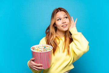 Teenager girl holding popcorns over isolated blue background listening to something by putting hand on the ear