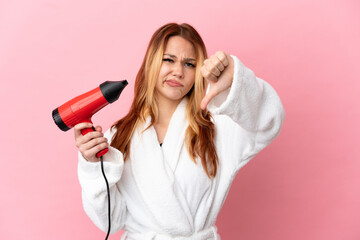 Teenager blonde girl holding a hairdryer over isolated pink background showing thumb down with negative expression