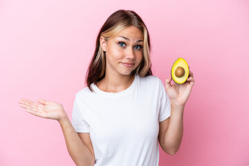 Young Russian woman holding avocado isolated on pink background having doubts while raising hands