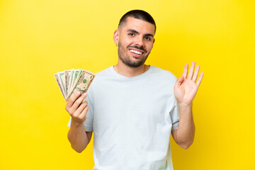 Young caucasian man taking a lot of money isolated on yellow background saluting with hand with happy expression