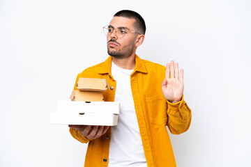Young caucasian man holding pizzas and burgers isolated on background making stop gesture and...