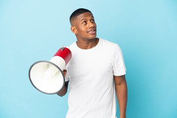 Young latin man isolated on blue background holding a megaphone and looking up while smiling