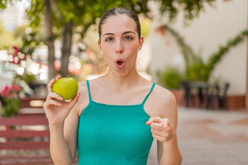 Young pretty woman with an apple at outdoors surprised and pointing front