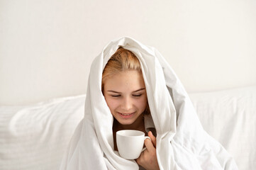 Sleepyhead, teenage girl wrapper in blanket sitting on bed with coffee cup, minimalist white...