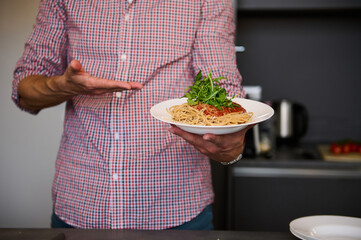 Close-up hands of young Italian chef showing white plate with Italian spaghetti capellini, standing in the home kitchen. Chef showing a served dish with Italian pasta. Italy. Food. Culture. Traditions
