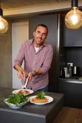 Caucasian handsome man 40 years old, grating cheese on freshly cooked Italian spaghetti with tomato sauce, smiling looking at camera while preparing family dinner at home kitchen. Italian cuisine