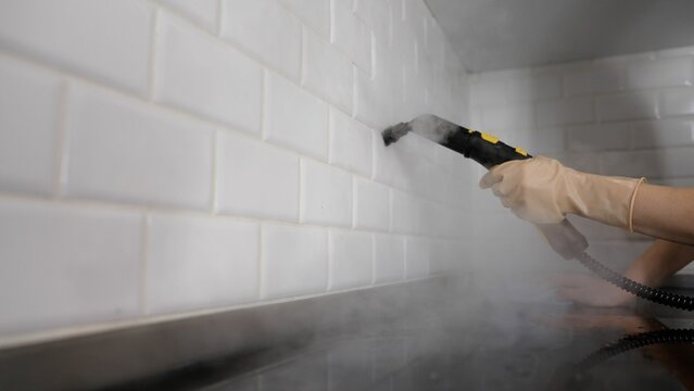 A person does the cleaning with hot water; a steam jet removes pressure dirt from the joints. The work of cleaning services Cleaning the black ceramic tile with a steam cleaner in the bathroom.