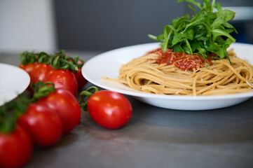 Still life with a white plate with Italian spaghetti capellini with tomato sauce, garnished with arugula green leaves and branch of red ripe organic tomatoes on the gray kitchen table