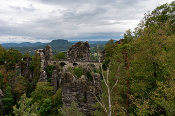 A mountain range with a bridge in the middle. The bridge is surrounded by trees and the mountains are covered in clouds