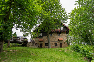 A stone house with a wooden porch sits in a lush green field. The house is surrounded by trees and has a bridge leading up to it. The scene is peaceful and serene