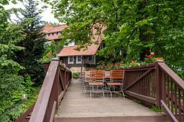 A wooden bridge with a house on the other side. There are several chairs and tables on the bridge, and potted plants are placed around the area. The scene gives off a peaceful and relaxing vibe