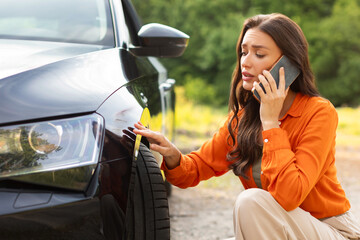 Sad young woman sitting near scratched auto and calling husband for help, having problem with new...