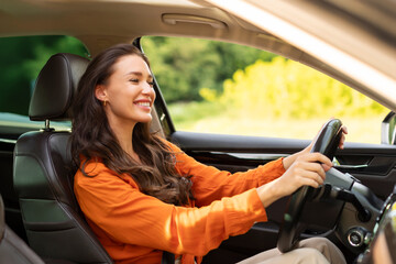 Happy cheerful young woman driver sitting in front seat, holding auto steering wheel, driving her...