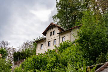 A large white house with a red roof sits on a hillside. The house is surrounded by trees and has a lot of windows. The sky is cloudy, giving the scene a somewhat somber mood