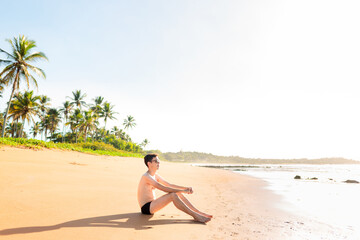 Brazilian man wearing a swimwear sitting on the beach sand to relax and enjoy the summer. He is looking at the sea. In the background, a sunny sky and some coconut trees.