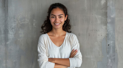 A young Latin woman with a warm smile stands in front of a gray wall. She has her arms crossed and is looking at the camera.