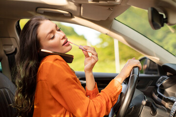 Busy female driver talking on cellphone and applying lipstick, while holding steering wheel,...