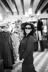 Obraz premium Photography in vintage style in black and white. A young girl tries on a hat in a store. Inside the boutique there is a characteristic atmosphere - a lot of clothes and accessories