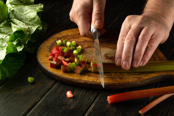 Male hands using a knife to cut rhubarb stalks on a wooden board for preparing a vegetarian dish at...