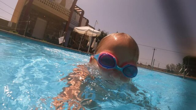 Close-up underwater view of a young girl with goggles swimming towards the camera in a vibrant blue pool