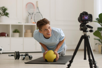 Smiling sports blogger training with medicine ball while recording fitness lesson with camera at...