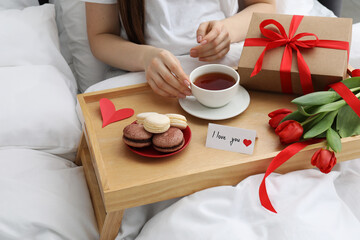 Tasty breakfast served in bed. Woman with tea, macarons, gift box, flowers and I Love You card at home, closeup