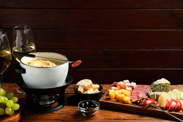Fork with piece of sausage, melted cheese in fondue pot and other products on wooden table. Space...
