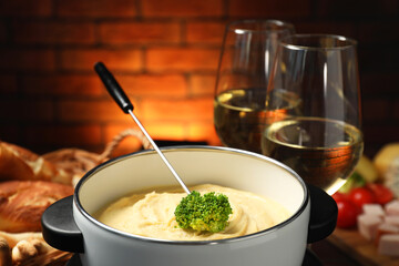 Fork with piece of broccoli, melted cheese in fondue pot, wine and snacks on blurred background,...