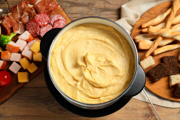Tasty melted cheese in fondue pot and snacks on wooden table, flat lay