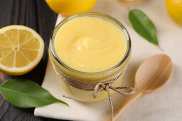 Delicious lemon curd in bowl, fresh citrus fruit, spoon and green leaves on wooden table, closeup