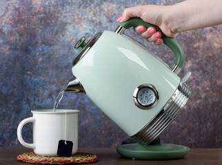 A woman pours hot water from a kettle into a cup with a disposable tea bag.