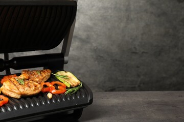 Electric grill with tasty meat, spices and vegetables on grey textured table, space for text